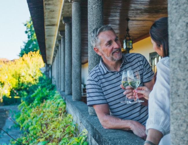 middle aged man and woman clinking classes of white wine in a portico overlooking a leafy garden