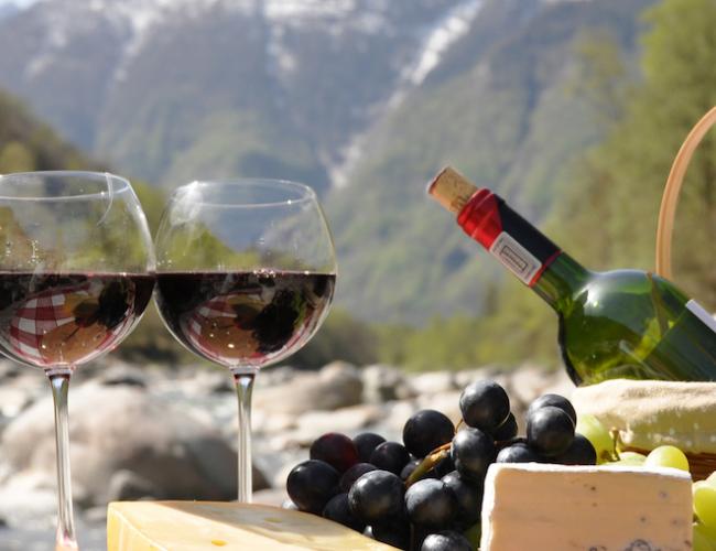 a picnic basket and two glasses of red wine, cheese and grapes in the foreground and a Swiss mountain scene in the background