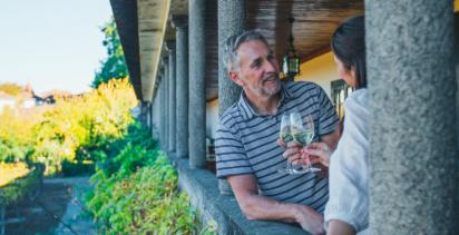 middle aged man and woman clinking classes of white wine in a portico overlooking a leafy garden