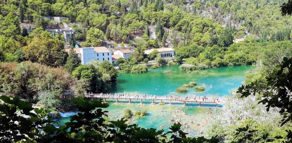 a bridge with people crossing and a lake with people swimming surrounded by verdant woodland, Krka National Park, Croatia